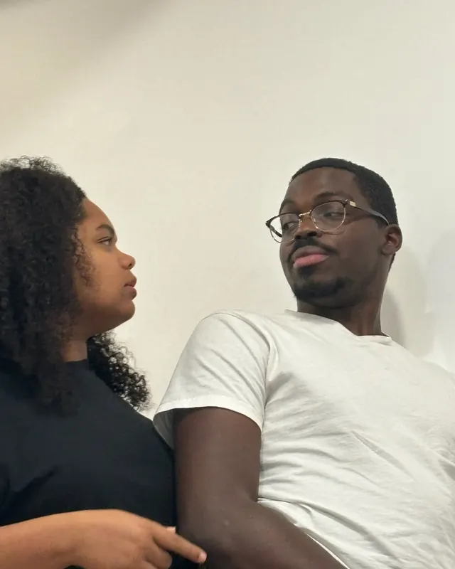 Nicole looks at Timothy, who gazes up and away, against a white wall.