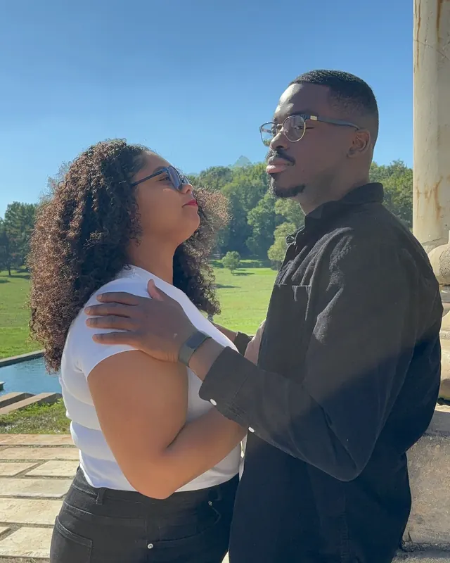 Nicole and Timothy embrace beside a stone column, smiling at each other under a blue sky.